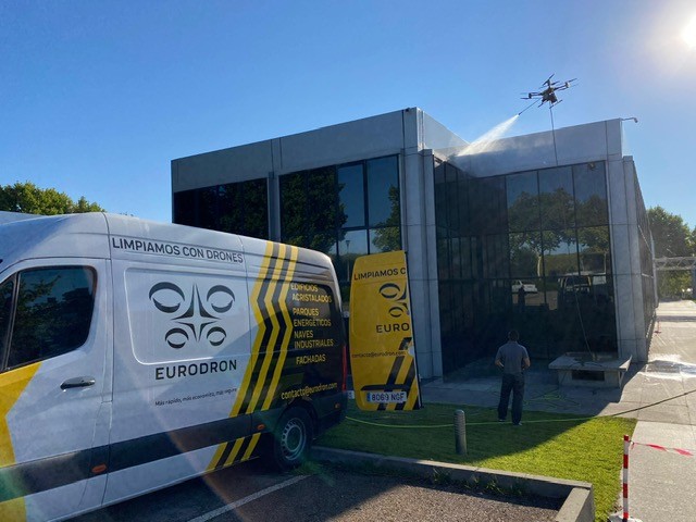 Furgoneta rotulada de EURODRON junto a un edificio de oficinas acristalado en Pozuelo de Alarcón mientras un dron pulveriza agua sobre la fachada y un operario controla la intervención desde tierra